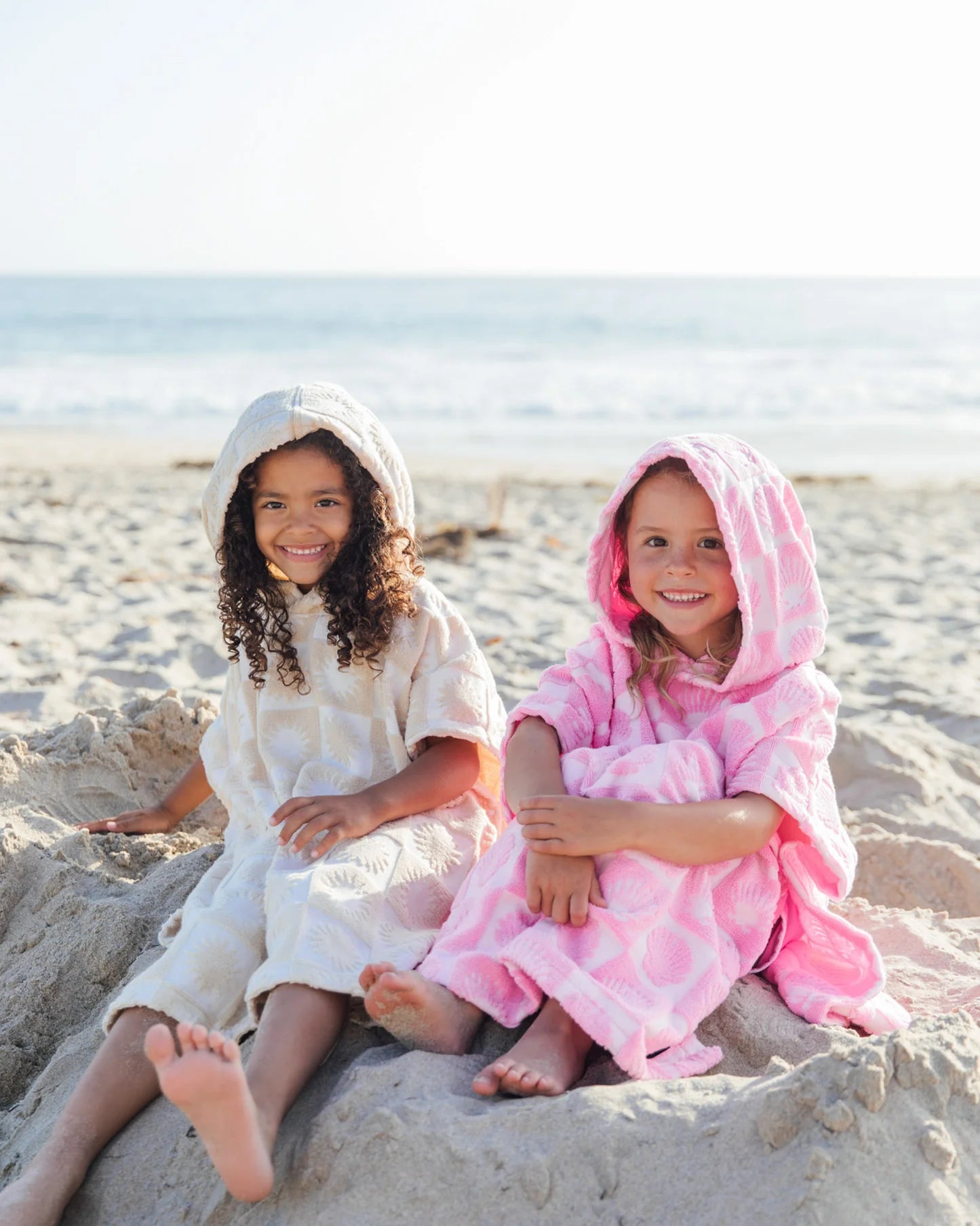 Two children sitting on a sandy beach wearing hooded towels.