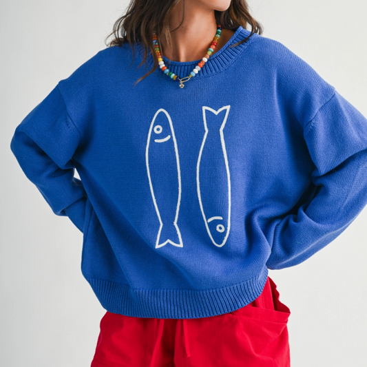 A person wearing a bright blue crewneck sweater with two white fish outlines on the front, paired with red pants and a colorful beaded necklace, against a light gray studio background.