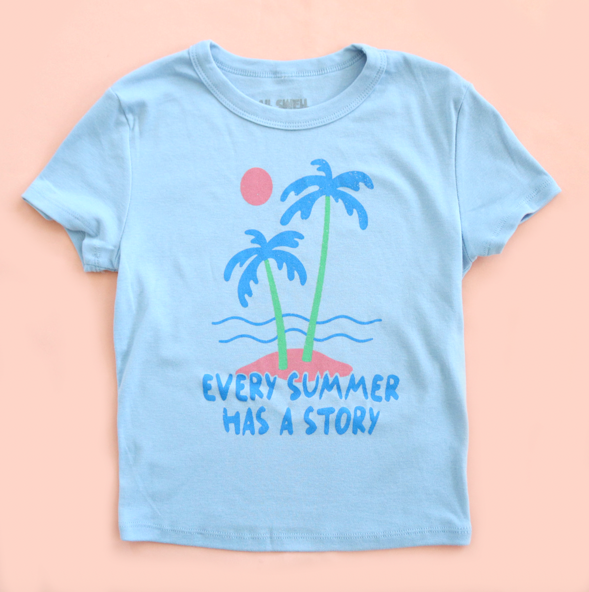 Light blue t-shirt with palm tree design and text on a peach background
