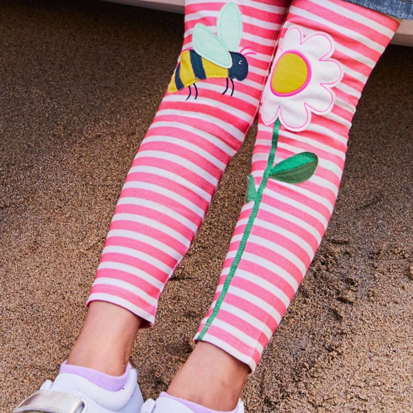 Pink and white striped leggings with a yellow and black bee applique on one leg and a white daisy with a green stem on the other, shown on a child's legs.