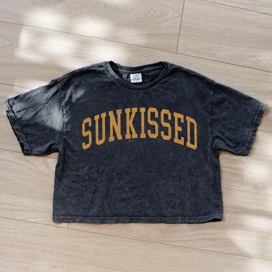 Black t-shirt with 'SUNKISSED' text on a wooden floor
