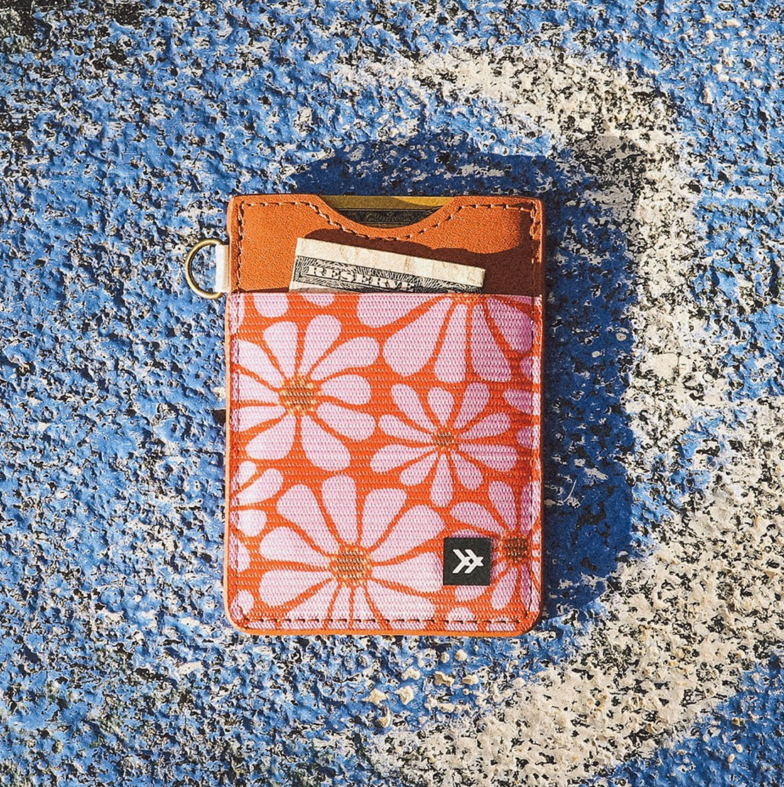 A slim card holder wallet with a retro orange and pink floral pattern, brown leather trim, and a small black logo patch, shown on a textured blue and white surface.