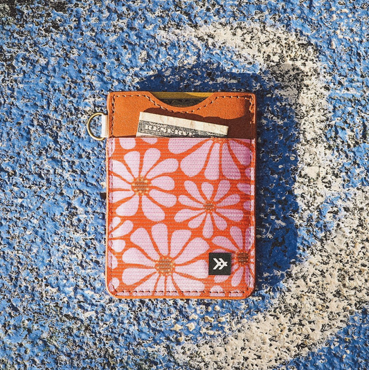 A slim card holder wallet with a retro orange and pink floral pattern, brown leather trim, and a small black logo patch, shown on a textured blue and white surface.